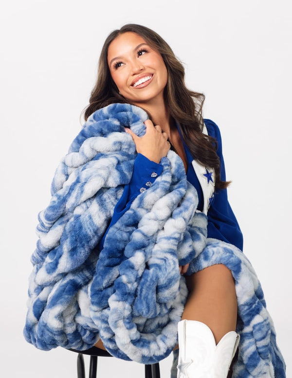 A smiling woman seated on a stool wrapped in a chunky, oversized blue and white marled throw blanket.