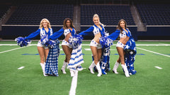 Four female cheerleaders in matching blue and white uniforms with pom-poms pose on a green football field in an indoor stadium with dark blue stadium seats in the background. Each of them are holding a different plush blanket.