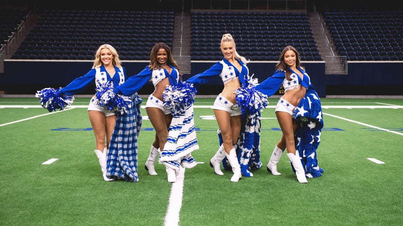 Four female cheerleaders in matching blue and white uniforms with pom-poms pose on a green football field in an indoor stadium with dark blue stadium seats in the background. Each of them are holding a different plush blanket.