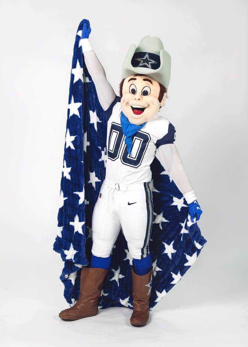 A Dallas Cowboys mascot character wearing a white and blue football uniform, gray cowboy hat, and blue blanket with white stars, holding the cape open against a light gray studio background.