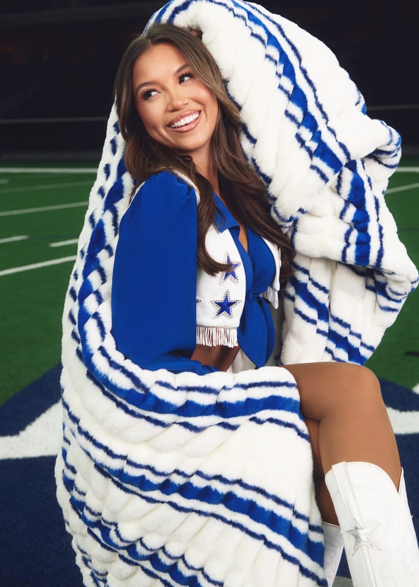 A woman wrapped in a large white and blue striped plush blanket, wearing a Dallas Cowboys cheerleader outfit on a football field.