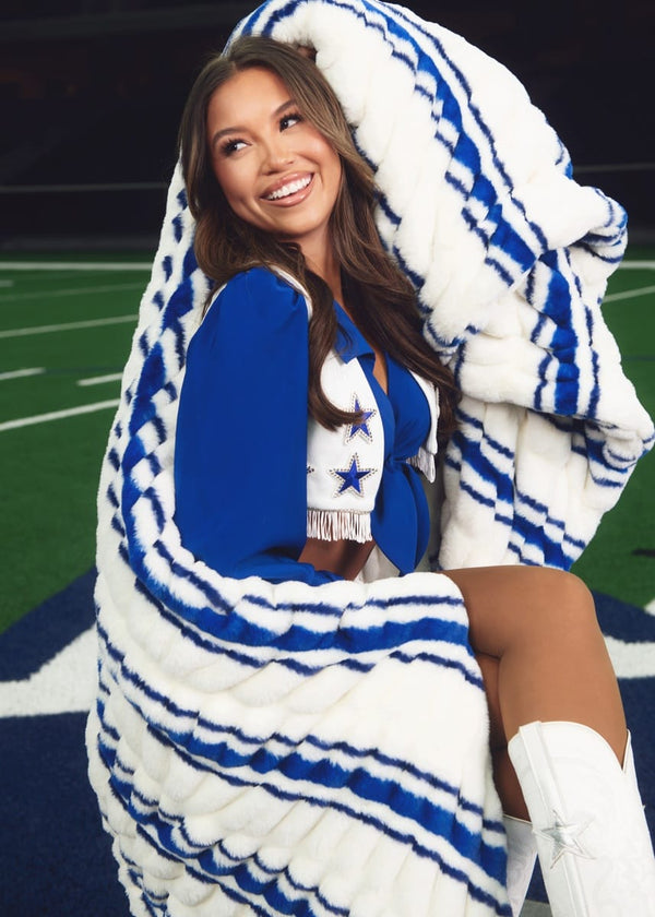 A woman wrapped in a large white and blue striped plush blanket, wearing a Dallas Cowboys cheerleader outfit on a football field.