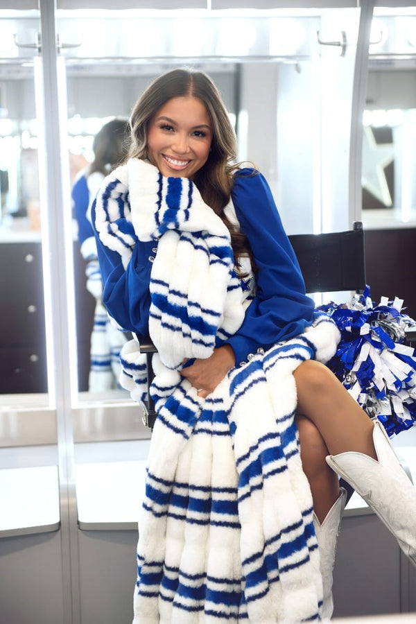 A woman wearing a plush faux fur blanket with blue and white stripes, styled with a matching blue sweatshirt and white cowboy boots in a modern indoor setting.