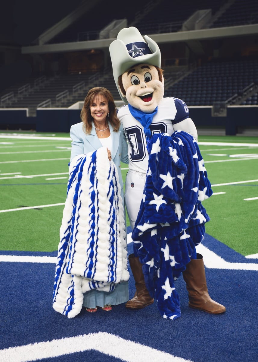 A woman and a Dallas Cowboys mascot pose together on a football field, each holding a blue and white star-patterned blanket. The woman wears a light blue blazer, and the mascot is dressed in a white and blue cowboy outfit with a cowboy hat.