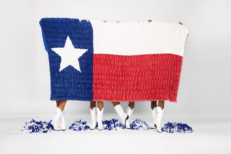 A large Texas flag blanket held up by three people standing in white cowboy boots, with blue and white pom-poms on the floor against a clean white studio background.