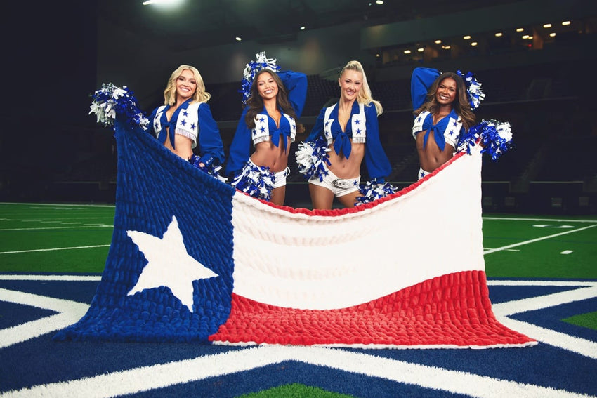 Four cheerleaders in matching blue and white uniforms pose on a football field holding a large, textured Texas flag blanket.