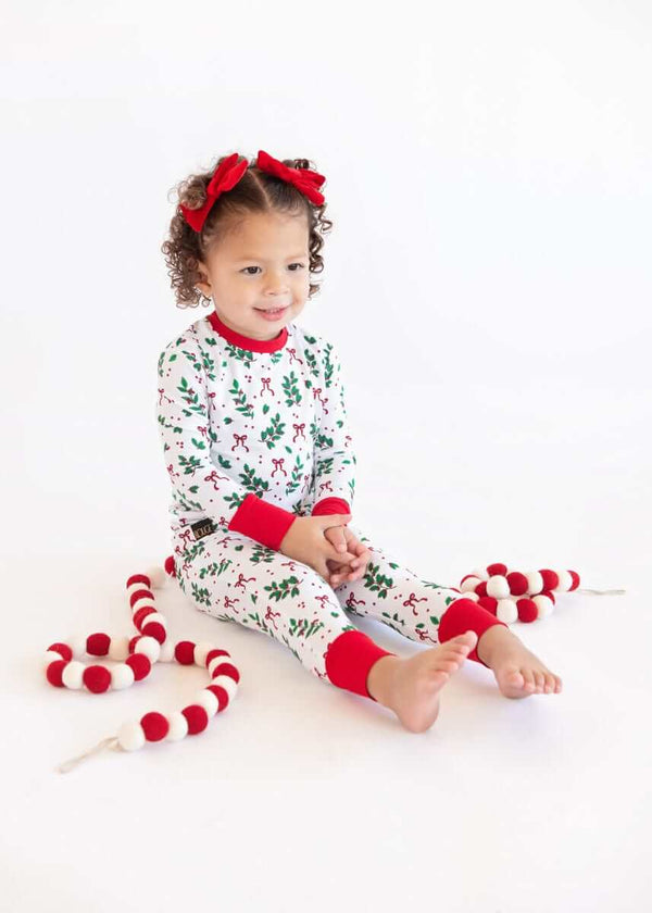 Toddler sitting on a white background wearing a Snug-Fitting Unisex Toddler Matching Set holiday sleeper with holly print and red cuffs.