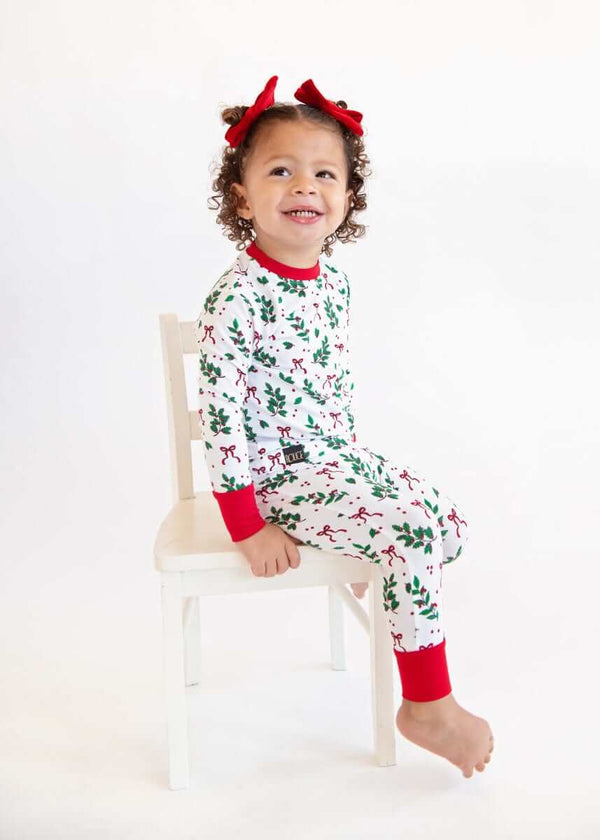 Toddler sitting on a chair wearing a Snug-Fitting Unisex Toddler Matching Set holiday sleeper with holly print and red cuffs.