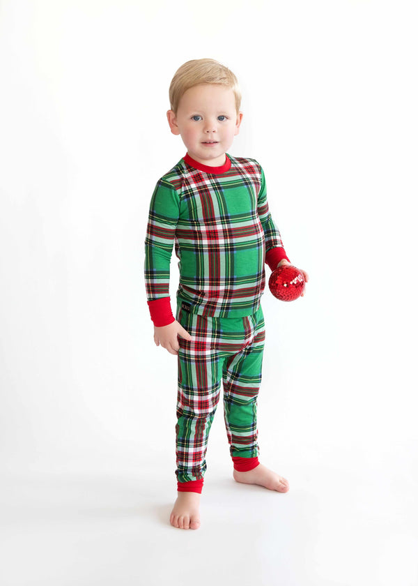 Toddler wearing green plaid holiday pajamas with red cuffs, standing barefoot and holding a small red ornament.