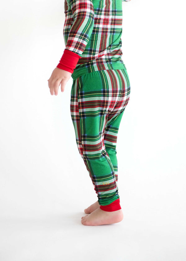 Side view of a toddler wearing green plaid holiday pajamas with red cuffs, standing barefoot against a white background.