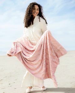 Woman in a flowing white dress with a pink blanket standing on sand with a blue sky background