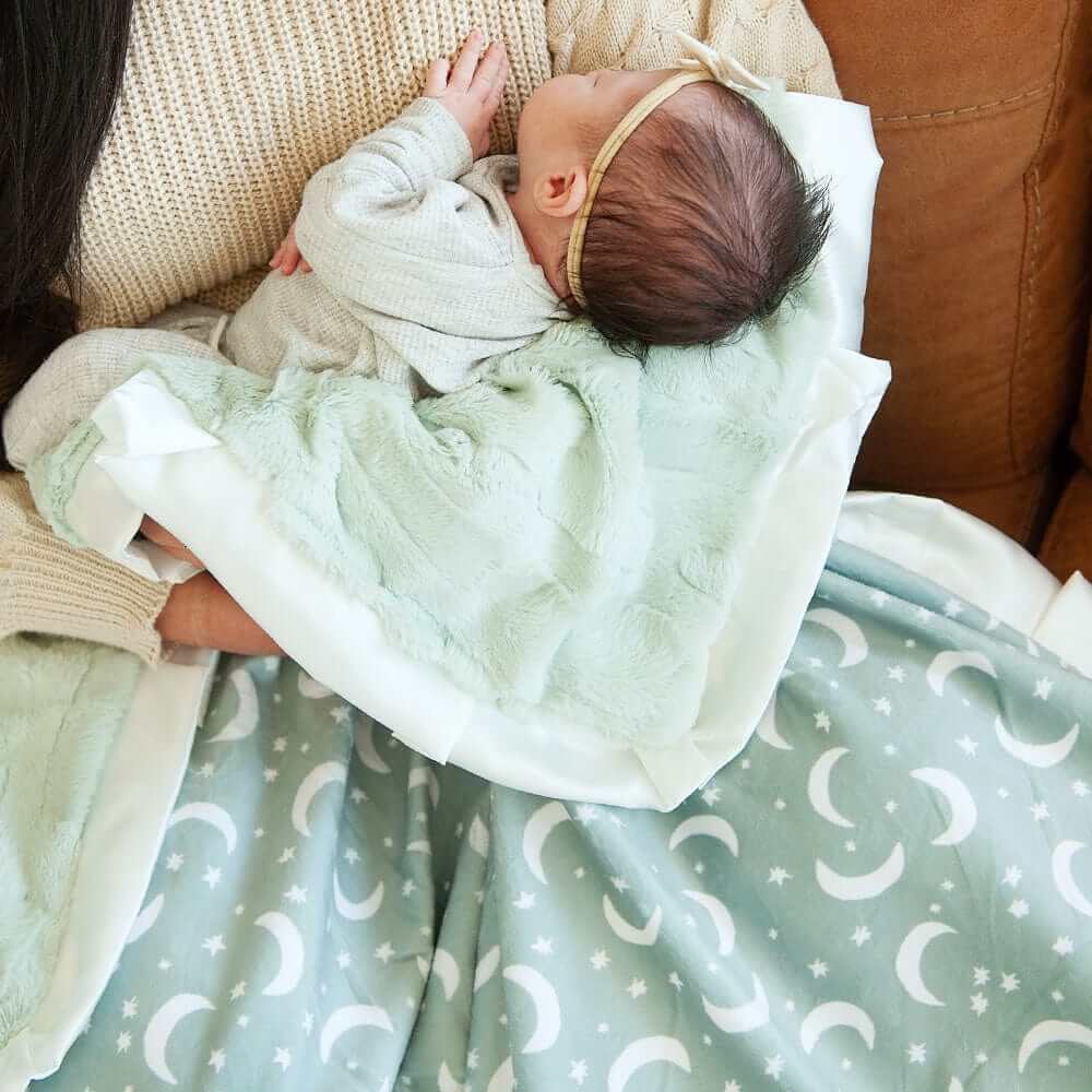 A cozy baby wrapped in a Twilight Sky Blanket featuring cream stars and moons on sage green fabric.