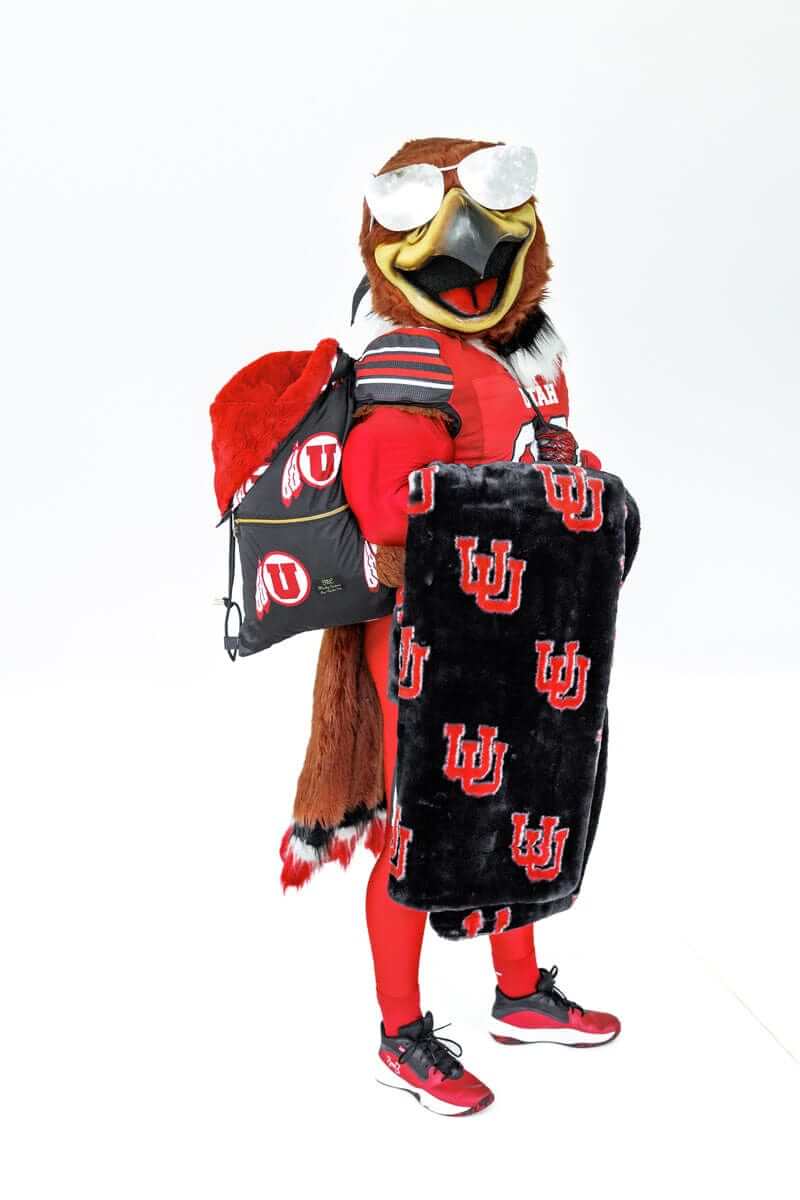 Mascot in red uniform holding black U logo  blanket and matching drawstring bag.