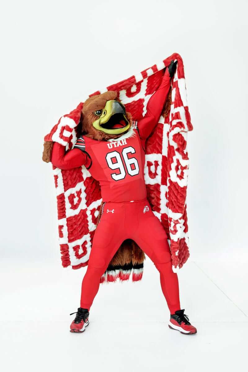 University of Utah mascot in red uniform holding red and white checkered blanket overhead in studio.