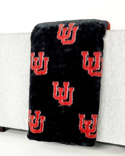 Black plush Minky Couture University of Utah blanket with red U logo draped over light gray sofa arm.