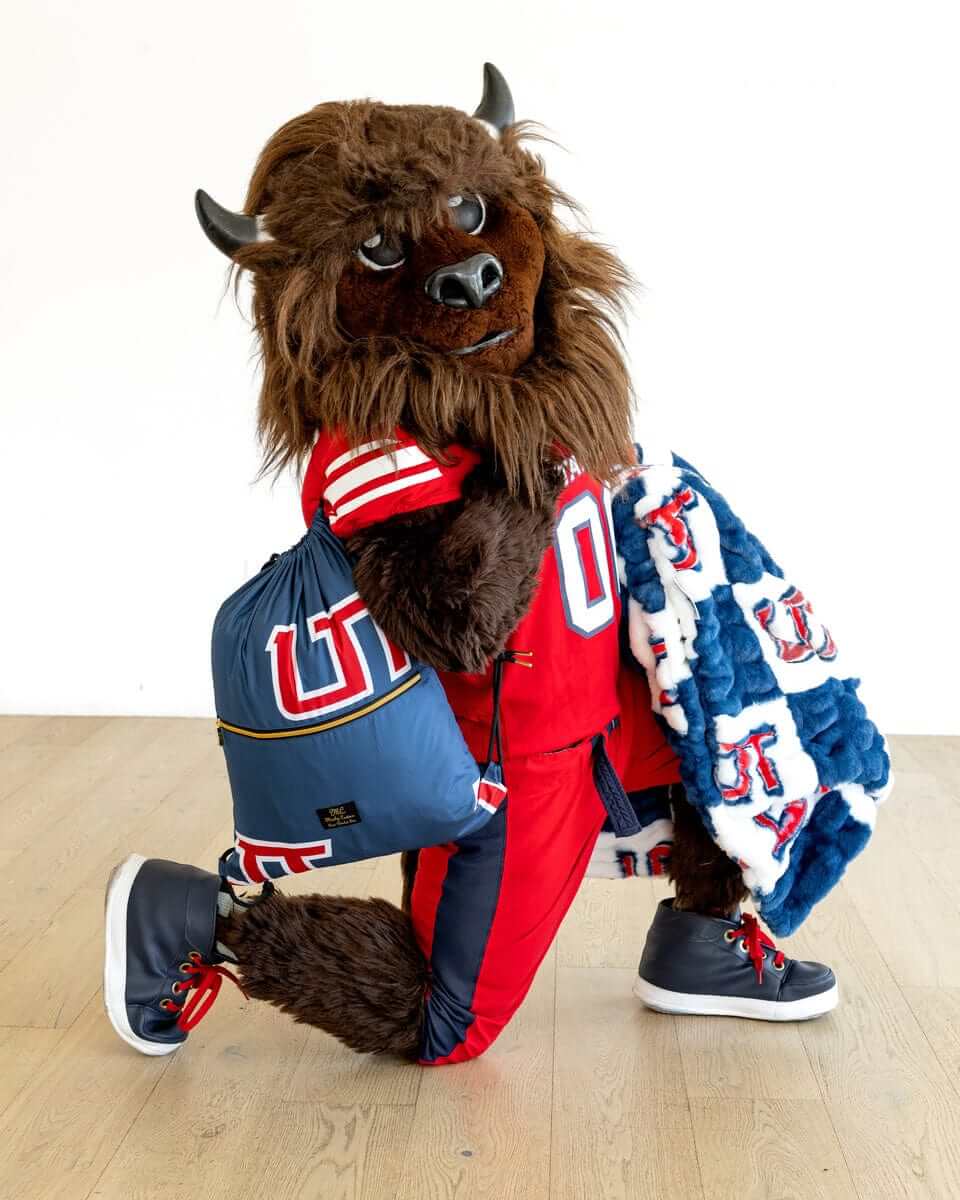 Utah Tech Trailblazers mascot kneeling with Minky Couture blue UT pillow and checkered team blanket indoors.