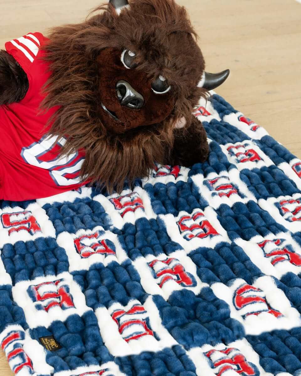 Buffalo mascot lying on blue and white Utah Tech Trailblazers checkered plush blanket indoors.