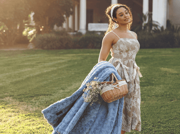 Young woman in floral dress holding blue blanket and basket on green grass, sunlight creating a warm atmosphere.