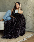 A woman sits on a white sofa wrapped in a plush, Ultra Sorbet Black Blanket, set against a textured gray concrete wall.