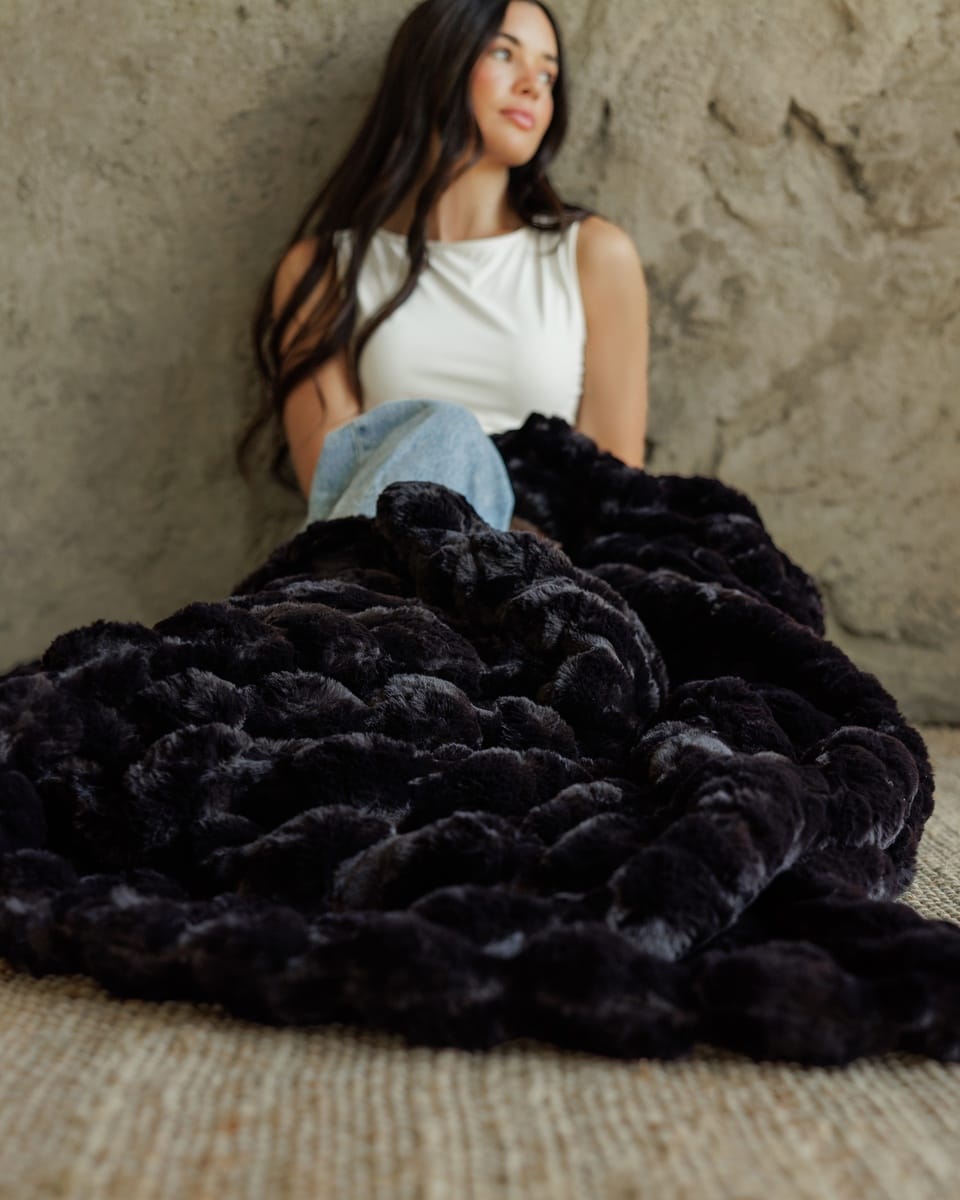A woman sits against a textured stone wall, wrapped in a chunky, oversized Ultra Sorbet Black Blanket. The blanket is a very dark plum or black color with a highly textured, boucle-like looped pattern.