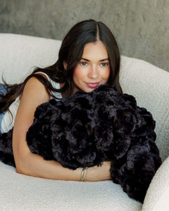 A woman with long dark hair reclines on a textured cream sofa, holding a Ultra Sorbet Black Blanket.