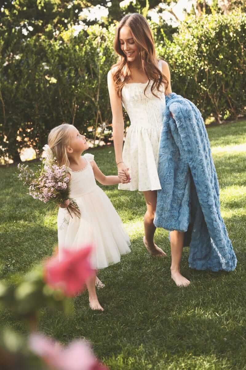 Mother and daughter in white dresses walking in a garden, with a cozy Ultra Sorbet Faded Denim Blanket draped over one arm.