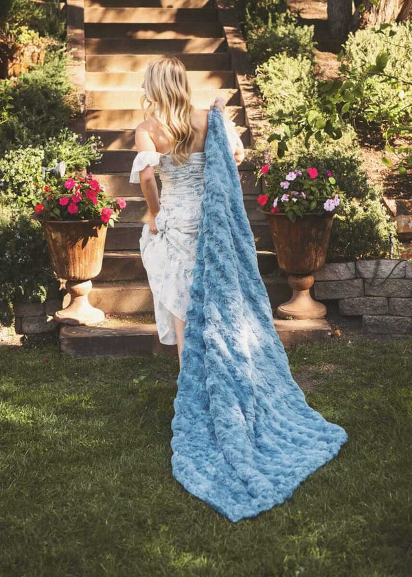 Woman draped in Ultra Sorbet Faded Denim Blanket walking through a garden with colorful flowers.