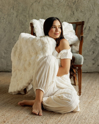 A woman sits barefoot on a woven rug in front of a rustic textured wall, wearing a white off-the-shoulder dress and harem-style pants. A large Ultra Sorbet Marshmallow Cream Blanket is draped over her and over the back of a wooden chair behind her.