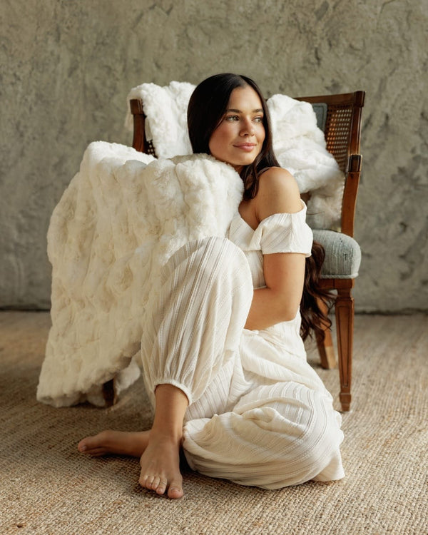 A woman sits barefoot on a woven rug in front of a rustic textured wall, wearing a white off-the-shoulder dress and harem-style pants. A large Ultra Sorbet Marshmallow Cream Blanket is draped over her and over the back of a wooden chair behind her.