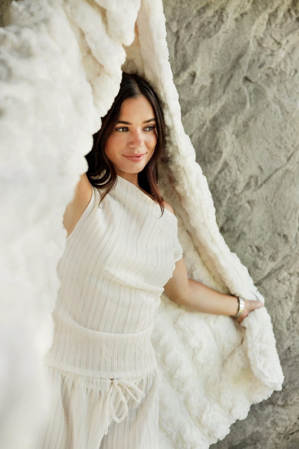 A woman wearing a white ribbed knit one-shoulder top and matching bottoms, posing with a chunky white Ultra Sorbet Marshmallow Cream Blanket against a textured gray stone wall.