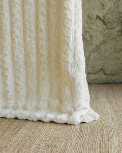 A close-up of a textured cream or off-white Ultra Sorbet Marshmallow Cream Blanket draped over a woven beige rug, with a stone wall in the background.