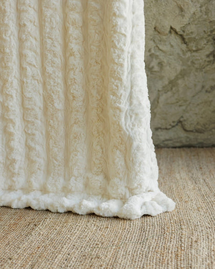 A close-up of a textured cream or off-white Ultra Sorbet Marshmallow Cream Blanket draped over a woven beige rug, with a stone wall in the background.