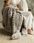 A cozy textured Ultra Sorbet Silver Cloud Blanket in a warm taupe-gray color is draped over a person sitting in a wooden chair. The person is wearing a cream-colored ribbed dress, and the scene is set on a woven jute rug against a neutral stone wall.