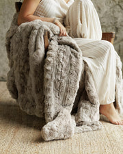 A cozy textured Ultra Sorbet Silver Cloud Blanket in a warm taupe-gray color is draped over a person sitting in a wooden chair. The person is wearing a cream-colored ribbed dress, and the scene is set on a woven jute rug against a neutral stone wall.