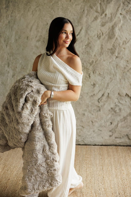 A woman wearing a white one-shoulder ribbed dress and holding a chunky light Ultra Sorbet Silver Cloud Blanket, standing against a textured beige wall.