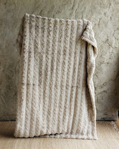 A chunky cable-knit Ultra Sorbet Silver Cloud Blanket, draped over a rustic stone wall and light wood floor.