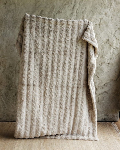 A chunky cable-knit Ultra Sorbet Silver Cloud Blanket, draped over a rustic stone wall and light wood floor.