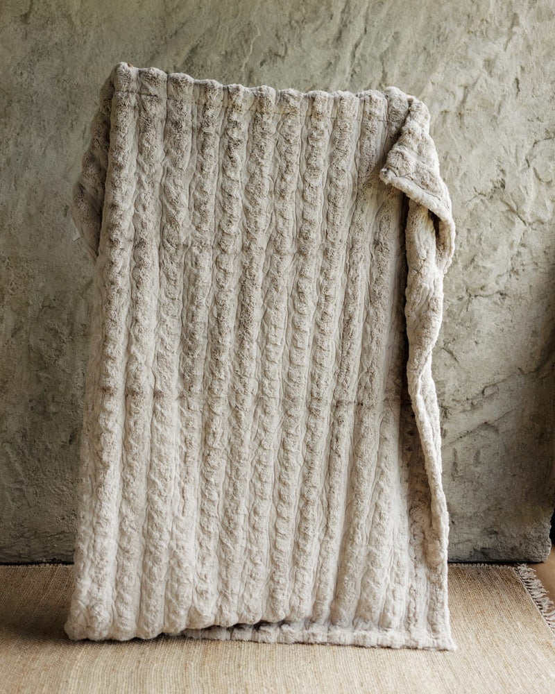 A chunky cable-knit Ultra Sorbet Silver Cloud Blanket, draped over a rustic stone wall and light wood floor.