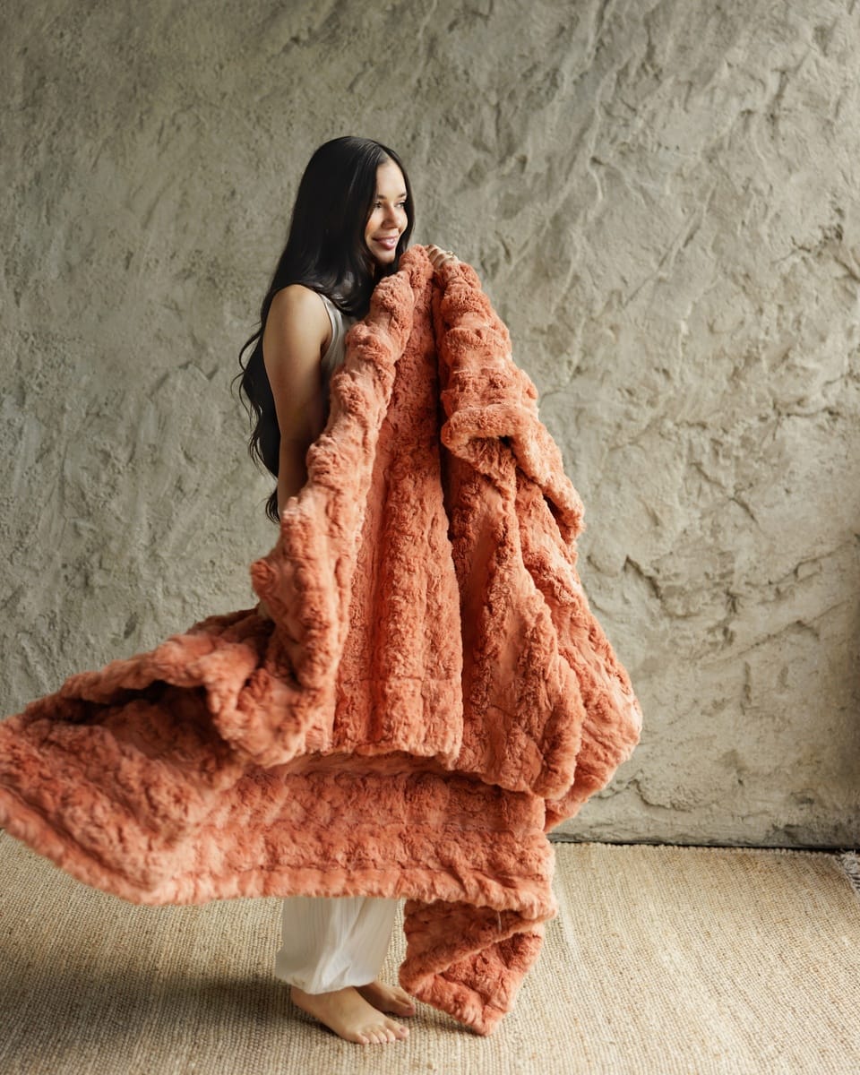 A woman stands barefoot against a textured beige wall, holding and draping a Ultra Sorbet Tawny Spice Blanket in a warm terracotta or burnt orange color.