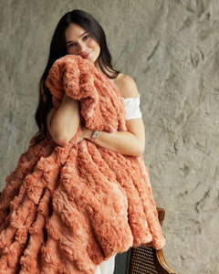 A woman with long dark hair holds a chunky, Ultra Sorbet Tawny Spice Blanket in front of her. She is wearing a white off-the-shoulder top and a silver watch, posed against a textured gray concrete wall.