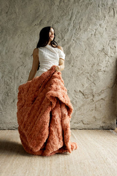 A woman stands in a room with a textured plaster wall and light wood floor, holding up a large, Ultra Sorbet Tawny Spice Blanket. She is wearing a white off-the-shoulder dress.