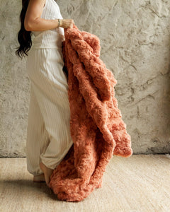A woman in a cream ribbed jumpsuit holds up a chunky, textured Ultra Sorbet Tawny Spice Blanket in front of a rustic concrete wall.