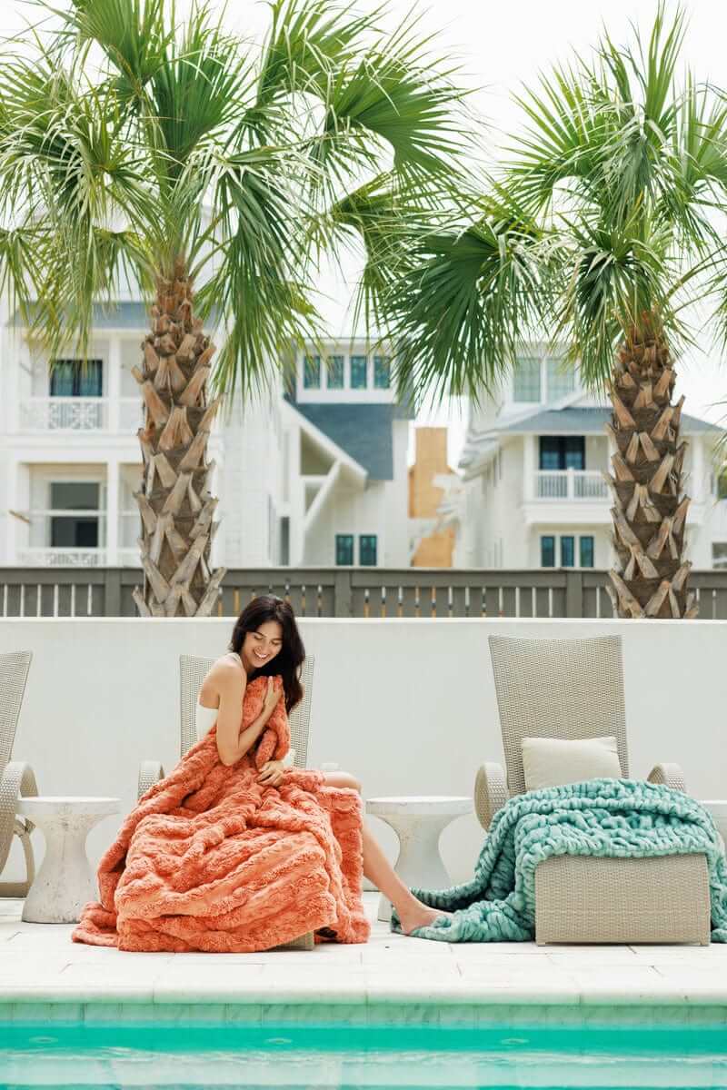 Woman relaxing by the pool with the Ultra Sorbet Tawny Spice Blanket and a teal blanket, surrounded by palm trees.