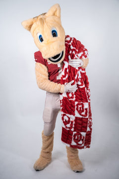 A University of Oklahoma Sooners mascot  holding a red and white plush Minky Couture blanket, photographed against a light gray studio background.