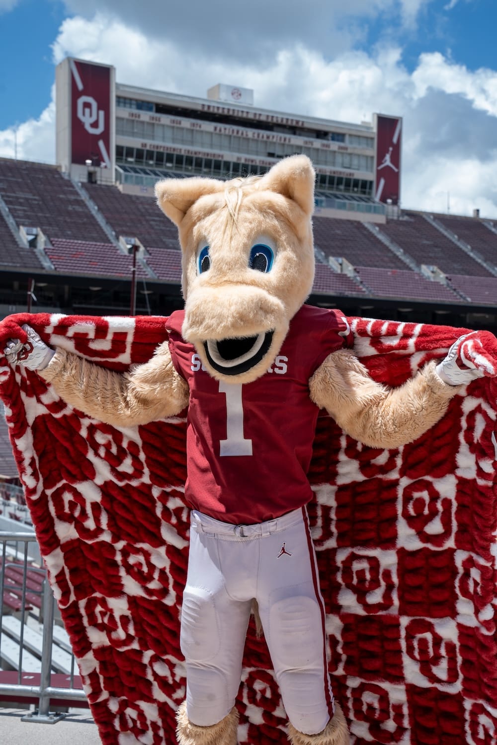 Oklahoma Sooners mascot character standing in a stadium, wearing a red jersey with the number 1 and white pants, holding up a hugs University of Oklahoma blanket. Stadium seating and sky are visible in the background.