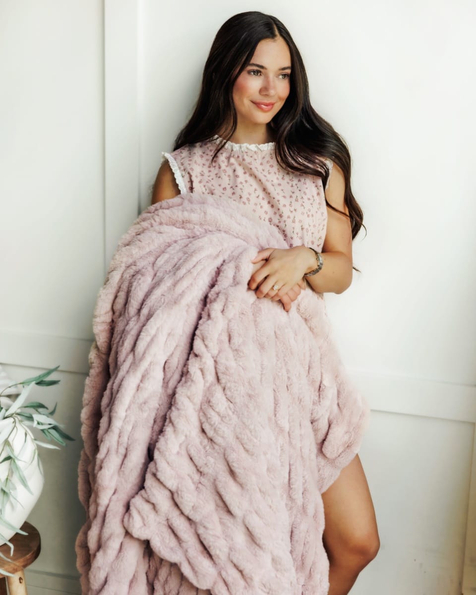 A woman with long dark hair is wrapped in a chunky, dusty pink plush blanket, wearing a light pink textured top. She is standing against a white paneled wall.