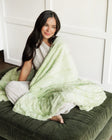A woman with long dark hair sits barefoot on a dark green velvet ottoman, wrapped in a Minky Couture Sorbet Lemon Lime plush throw blanket. She is wearing a white ribbed top and white pants, set against a clean white paneled wall.