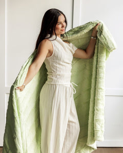 A woman in a cream pleated jumpsuit holds up a large, textured Minky Couture Sorbet Lemon Lime blanket with both hands in a brightly lit room with white paneled walls.