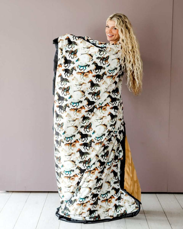 A woman smiling while holding a Stallion Blanket featuring watercolor horses on a cream background with a black satin ruffle.