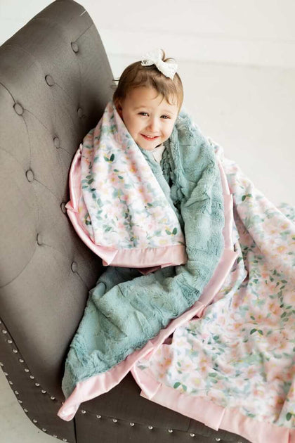 Little girl sitting on a chair wrapped up in a blush and blue floral patterned Minky Couture blanket.